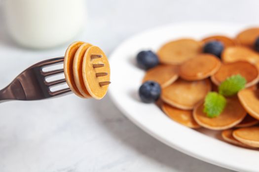 Trendy homemade breakfast, pancake cereal, mini pancakes on white plate on white concrete background with fork and blueberry, leaves of lemon balm and peppermint and bottle of milk