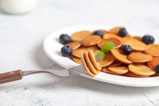 Trendy homemade breakfast, pancake cereal, mini pancakes on white plate on white concrete background with fork and blueberry, leaves of lemon balm and peppermint and bottle of milk