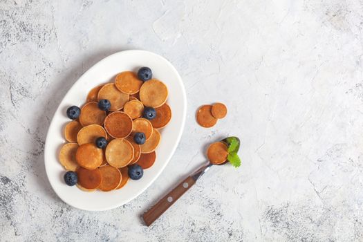 Trendy homemade breakfast, pancake cereal, mini pancakes on white plate on white concrete background with spoon and blueberry, leaves of lemon balm and peppermint