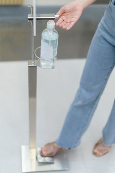 Woman using sanitiser dispenser foot pump. Hand free alcohol hand gel dispenser. Stop Coronavirus (Covid-19) Spread.