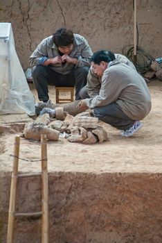 Xian, China - May 1, 2010: Terracotta Army excavation site. 3 researchers squat on dirt near trench with ladder and discuss found pieces of sculptures.