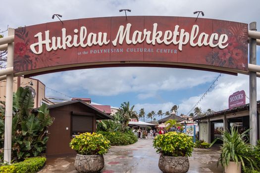 Laie, Oahu, Hawaii, USA. - January 09, 2020: Polynesian Cultural Center. Welcome sign at entrance to Hukilau Marketplace under blue cloudscape. One-story buildings, people and green vegetation.