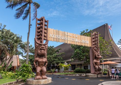Laie, Oahu, Hawaii, USA. - January 09, 2020: Polynesian Cultural Center. Monumental gate into the park with brown giant aboriginal statues. People and green vegetation under blue sky.
