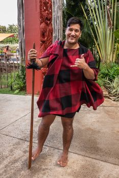 Laie, Oahu, Hawaii, USA. - January 09, 2020: Polynesian Cultural Center. Closeup of young male Maori warrior showing the Shaka sign. Black and red garb. Back is green vegetation and maroon totem sculptures.