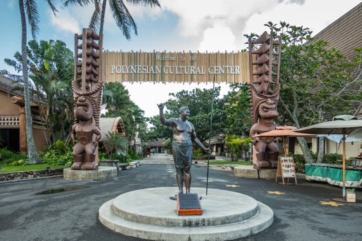 Laie, Oahu, Hawaii, USA. - January 09, 2020: Polynesian Cultural Center. Bronze statue of Hamana Kalili giving the Shaka sign in front of monumental entrance gate to the park. Green foliage, blue cloudscape.