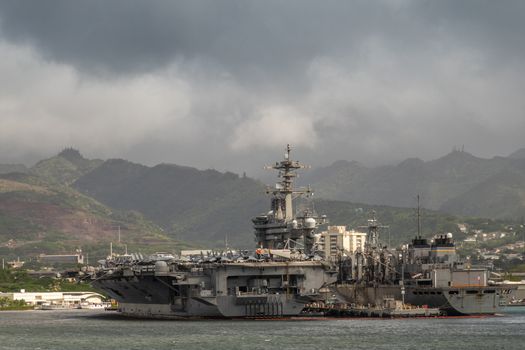 Oahu, Hawaii, USA. - January 10, 2020: Pearl Harbor. Stern sides of Gray Abraham Lincoln aircraft carrier docked with USNS Arctic under dark raining cloudscape on top of gray water. Mountains on horizon.