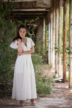 Beautiful young woman wears a long white dress while posing in the outdoors of a ruined environment