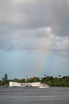 Oahu, Hawaii, USA. - January 10, 2020: Pearl Harbor. Portrait of White USS Arizona Memorial with rainbow landing on it. Green belt of foliage in back separating gray sea and light blue cloudscape.
