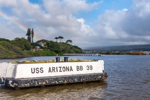 Oahu, Hawaii, USA. - January 10, 2020: Pearl Harbor. Historic white off-dock mooring point for USS Arizona on blueish water under blue cloudscape. Belt of foliage and rainbow.