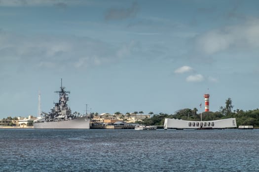 Oahu, Hawaii, USA. - January 10, 2020: Pearl Harbor. Gray USS Missouri together with USS Arizona Memorial side by side on blueish water and under blue cloudscape. Airfield tower, Green foliage belt.