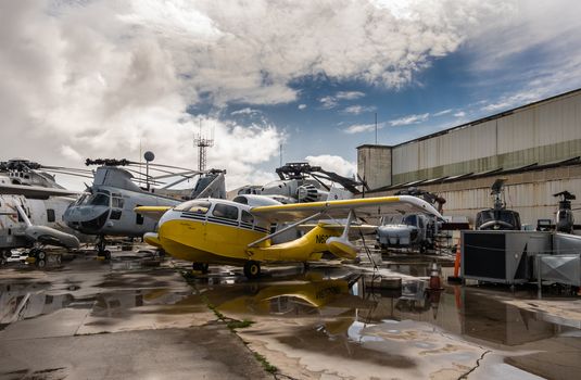 Oahu, Hawaii, USA. - January 10, 2020: Pearl Harbor Aviation Museum. Group of helicopters and yellow aircraft outside hangar under blue cloudscape on wet tarmac.