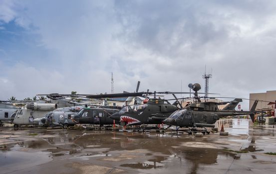 Oahu, Hawaii, USA. - January 10, 2020: Pearl Harbor Aviation Museum. Group of different old helicopters on wet tarmac outside hangar under cloudscape.