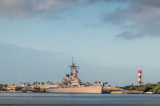 Oahu, Hawaii, USA. - January 10, 2020: Pearl Harbor. USS Missouri Battle Ship docked on blue water and under bly sky. Sun falls on gray hull. Red-white control tower of airport.