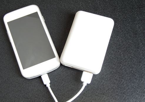 A white phone with a black screen and a copy of space is charged using a white power Bank. Top view, black background.