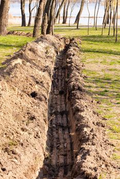 A trench in the ground for laying a pipe, or tubes, in the park, under the trees, close to the river, with spring light