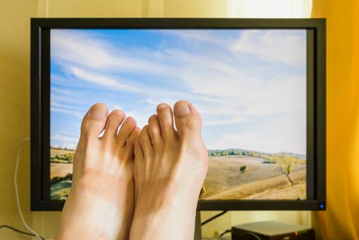 Man's feet in front of a computer monitor with a nice sunny italian landscape with a cloudy sky to symbolize the wish of holidays rest
