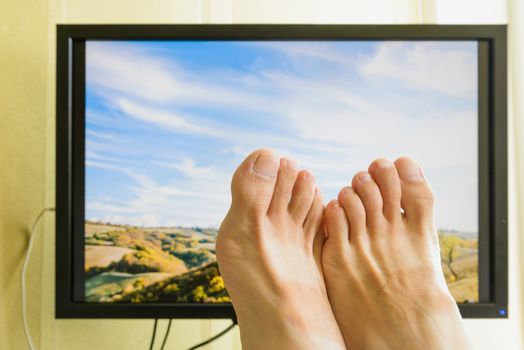 Man's feet in front of a computer monitor with a nice sunny italian landscape with a cloudy sky to symbolize the wish of holidays rest