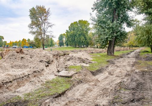 A trench in the sandy ground for laying a pipe in the park, under the trees, with evening autumn light