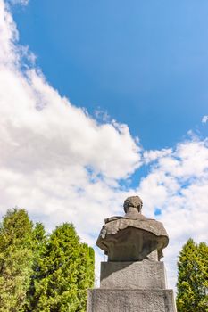 Statue bust of an unknown man, surrounded by trees, seen from back against a blue sky