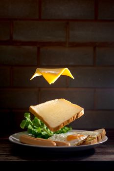 Food and breakfast levitation concept. Sliced bread and cheese float on sausage, fried egg, and green lettuce in white plate on wooden table in the home kitchen. 