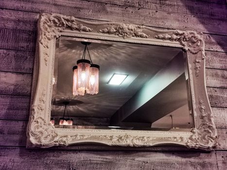 Mirror with vintage style frame hanging off the wooden wall with lighting in the reflections of a restaurant or hotel