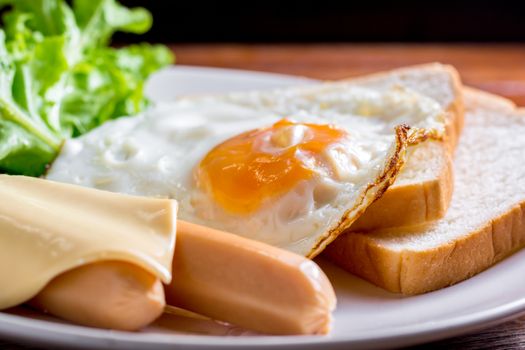 American breakfast concept. Fried egg with sliced bread, boiled chicken sausage, cheddar cheese, and fresh lettuce on a white plate in the home kitchen. Homemade food in the morning.