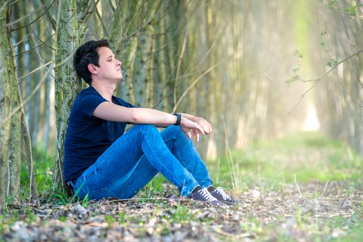 young man sitting by a tree in the woods thinking about life and resting.