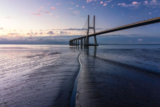 Vasco da Gama bridge at sunrise in Lisbon, Portugal. Vasco da Gama Bridge is a cable-stayed bridge flanked by viaducts and rangeviews that spans the Tagus River in Parque das Nações in Lisbon. 