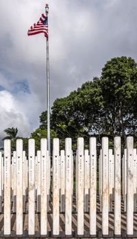 Oahu, Hawaii, USA. - January 10, 2020: Pearl Harbor, Closeup of group of white name-sticks and USA flag at USS Oklahoma memorial. Green foliage in back. Dark cloudscape.