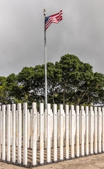 Oahu, Hawaii, USA. - January 10, 2020: Pearl Harbor, Closeup of group of white name-sticks and USA flag at corner on USS Oklahoma memorial. Green foliage in back. Dark cloudscape.