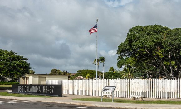 Oahu, Hawaii, USA. - January 10, 2020: Pearl Harbor, USS Oklahoma memorial. Black marble wall with the name in front of field of white marble sticks. Green foliage, US flag and dark cloudscape.