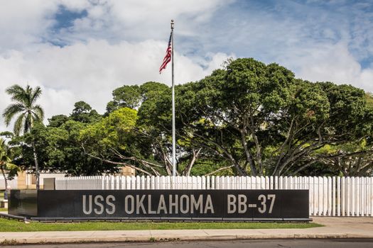 Oahu, Hawaii, USA. - January 10, 2020: Pearl Harbor, USS Oklahoma memorial. Black marble wall with the name in front of field of white marble sticks. Green foliage, US flag and blue cloudscape.