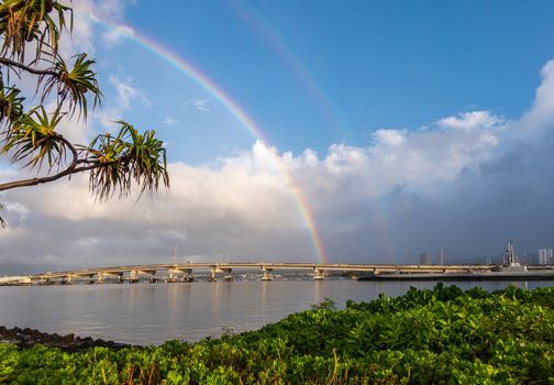 Oahu, Hawaii, USA. - January 10, 2020: Pearl Harbor. Ford Island bridge toubhed by double rainbow under blue sky with heavy rain clouds. Green foliage up front. Gray water.