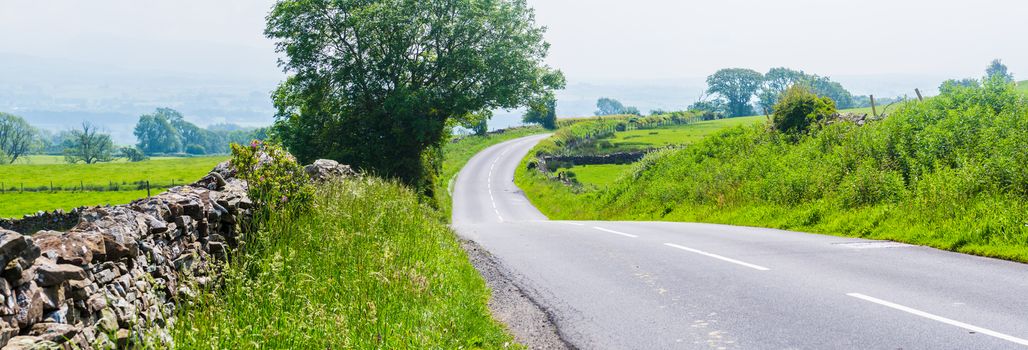 Panorama of a curved empty country road with dry stone walls and trees