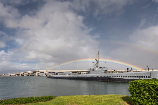 Oahu, Hawaii, USA. - January 10, 2020: Pearl Harbor. Rainbow straddles long submarine USS Bowfin sticking out of gray water under blue cloudscape. Ford Island bridge lines up with boat. Green foliage up front.