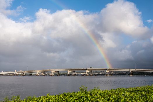 Oahu, Hawaii, USA. - January 10, 2020: Pearl Harbor. Ford Island bridge toubhed by rainbow under blue sky with heavy rain clouds. Green foliage up front. Gray water.