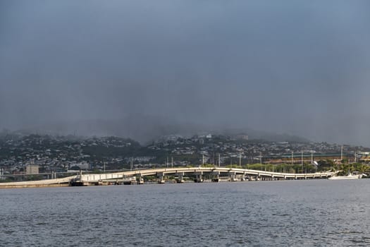 Oahu, Hawaii, USA. - January 10, 2020: Pearl Harbor. Ford Island bridge froms white line between gray water a belt of green hills with white houses. All under a dark foggy rain-filled sky.