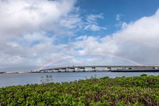 Oahu, Hawaii, USA. - January 10, 2020: Pearl Harbor. Rainbow straddles Ford Island bridge under blue coudscape/ Green foliage in front.