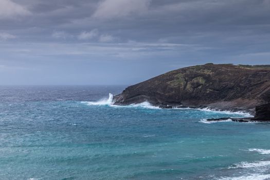 Oahu, Hawaii, USA. - January 11, 2020: Hanauma Bay Nature Preserve. White crashing waves of azure water on face of baboon cliff rock under heavy rain cloudscape.