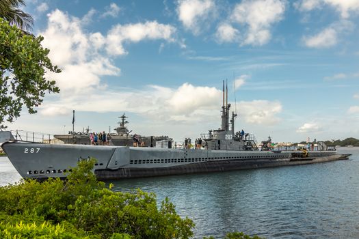 Oahu, Hawaii, USA. - January 10, 2020: Pearl Harbor. Long submarine USS Bowfin sticking out of blueish water under blue cloudscape. People on top. Deck of Abraham Lincoln aircraft carrier in back.