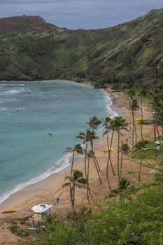 Oahu, Hawaii, USA. - January 11, 2020: Hanauma Bay Nature Preserve. Portrait of Brown sandy beach with palmtrees, white surf on azure water, green cliffs and rocks. All under heavy storm cloudscape.