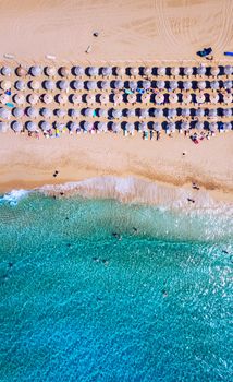 Aerial view of sandy beach with colorful umbrellas, swimming people in sea bay with transparent blue water at sunset in summer. Aerial top view on the beach, umbrellas, sand and sea waves.