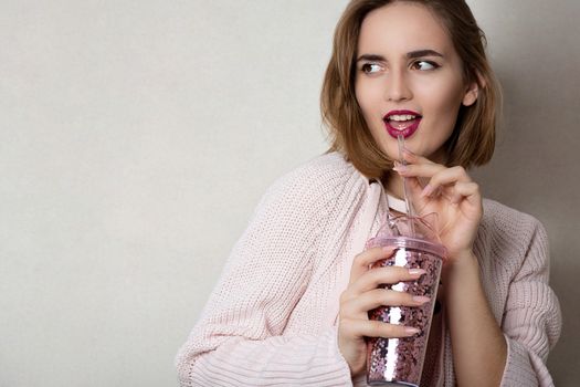 Fashionable young woman wears pink sweater holding glass of orange juice over a grey wall. Space for text