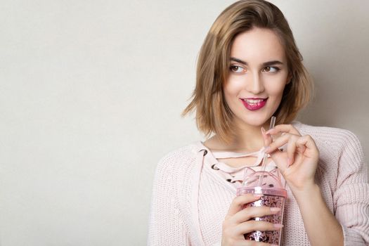 Seductive young woman wears pink sweater holding glass of orange juice over a grey wall. Space for text
