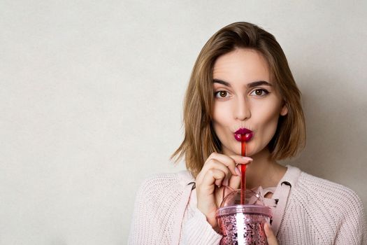 Flirty young woman wears pink sweater holding glass of orange juice over a grey wall. Space for text