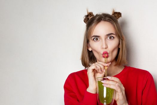 Funny young woman wears red sweater drinking kiwi juice over a grey background. Empty space