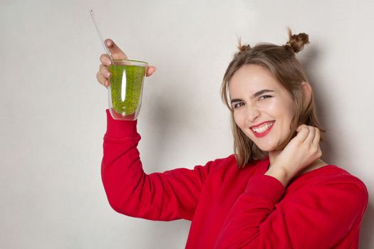 Cheerful young woman wears red sweater holding glass of kiwi juice over a light grey background. Empty space