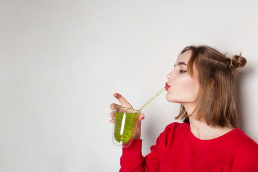 Lovely young woman wears red sweater drinking kiwi juice over a grey background. Empty space