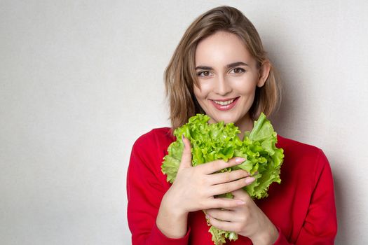 Attractive brunette woman wears red sweater holding lettuce over a grey background. Empty space