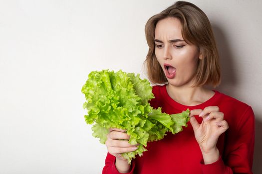 Worried brunette woman wears red sweater holding lettuce over a grey background. Empty space